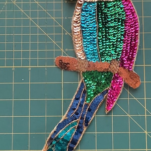 May include: A colorful parrot applique with a brown branch. The parrot features a red head, gold beaded beak, and body with teal, green, and pink sequins. The tail is a gradient of teal and blue sequins, with gold thread outlining the edges.
