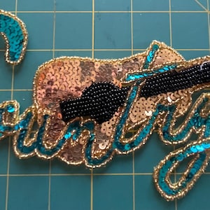 May include: A sequined patch featuring a guitar and the word "Country" in teal script. The guitar is made of gold sequins with a black beaded neck. The word "Country" is outlined in gold and teal sequins.
