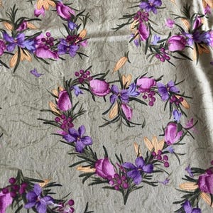 Peut inclure: Un tissu avec un motif floral répétitif. Le motif présente des arrangements en forme de cœur de fleurs violettes et roses, de feuilles vertes et d'accents orange sur un fond vert clair. Le tissu a un aspect texturé.