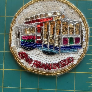 May include: Embroidered patch featuring a San Francisco cable car design. The patch is circular, with a white sequined background and a gold beaded border. The cable car is detailed with red, gold, and blue beads, and the text "SAN FRANCISCO" is embroidered in red.