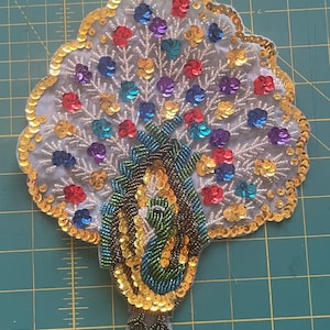 May include: A decorative peacock applique with a fan of colorful sequins in red, blue, purple, and gold. The peacock's body is detailed with green and gold beads, and the edges are trimmed with gold sequins.