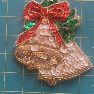 May include: A Christmas bell applique with a red sequined bow and green holly leaves. The bells are adorned with gold and white sequins, and outlined with gold thread. This festive embellishment is perfect for holiday crafts.
