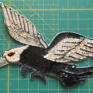 Può includere: Applicazione di aquila con perline e paillettes in volo. Le ali sono argentate, il corpo è nero e la testa è bianca con un occhio rosso. L'applicazione è su uno sfondo a griglia verde.