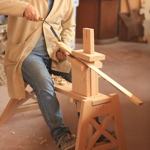 May include: A person using a wooden woodworking tool to shape a piece of wood. The tool is made of wood and has a simple design. The person is sitting on a wooden stool and is using a hand tool to shape the wood.
