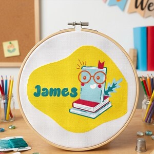 Op de afbeelding: Een ronde houten borduurring met witte stof en een kruissteekontwerp. Het ontwerp toont een gele vlek, een stapel boeken, een boek met een oranje bril en de naam James in blauwgroene cursieve letters op de witte stof.