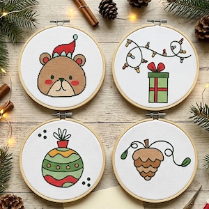 Op de afbeelding: Vier ronde houten borduurringen met kruissteekontwerpen op witte stof. De ontwerpen tonen een bruine beer met een rode muts, een groen cadeau met kerstverlichting, een kleurrijke gestreepte kerstbal en een bruine dennenappel.