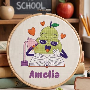 Könnte beinhalten: Ein runder Holzstickrahmen mit einem Kreuzstichmotiv einer grünen Birnenfigur, die unter einer rosa Schreibtischlampe in einem offenen Buch liest. Die Birne hat einen violetten Mund und dunkle Augen, umgeben von drei orangefarbenen Herzen. Der Name Amelia ist unten in violetten Buchstaben gestickt.