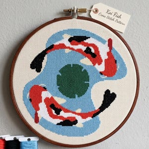 May include: A circular wooden embroidery hoop containing a cross stitch pattern of two koi fish swimming in a blue pond with a green lily pad. The fish are white, red, and black. A paper tag attached to the top reads Koi Fish Cross Stitch Pattern.