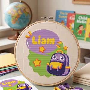 Puede incluir: Un bastidor de bordado circular de madera que contiene un diseño de punto de cruz sobre tela beige. El diseño presenta un globo de texto morado con el texto amarillo Liam, rodeado de estrellas amarillas y una mochila morada.