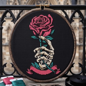 May include: A circular wooden embroidery hoop containing a cross-stitch design on black fabric. The design features a skeletal hand holding a single pink rose with green leaves, positioned above a pink banner at the bottom of the hoop.