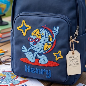 Puede incluir: Una mochila azul marino con un diseño de punto de cruz de un globo terráqueo con gafas de sol rojas haciendo el signo de la paz. El diseño incluye estrellas amarillas y el nombre Henry. Una etiqueta dice BACK-TO-SCHOOL CUSTOM STITCH KIT.