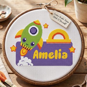 Puede incluir: Un bastidor de bordado circular de madera con tela blanca y un diseño de punto de cruz de un cohete verde, un arcoíris amarillo y estrellas naranjas. Una banda morada muestra el texto amarillo Amelia. Una etiqueta dice Custom Amelia Design.