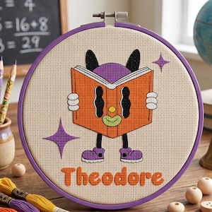 May include: A circular purple embroidery hoop containing a cross-stitch design of a character with black ears, a yellow nose, and a green mouth holding an orange book. The name Theodore is stitched in orange below the character with purple star accents.