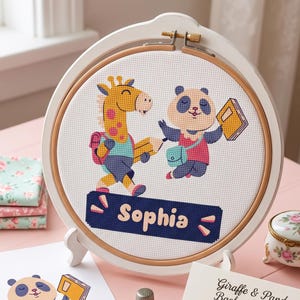 Puede incluir: Un bastidor de bordado circular de madera con tela Aida blanca y un diseño de punto de cruz de una jirafa amarilla y un panda blanco y negro con material escolar. Debajo de los animales hay una banda azul oscuro con el nombre Sophia en amarillo.