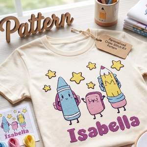 Puede incluir: Una camiseta de color crema con un diseño de punto de cruz de un crayón azul, una goma de borrar rosa y un lápiz amarillo con caras sonrientes, rodeados de estrellas amarillas. El nombre Isabella está bordado en morado bajo los personajes.