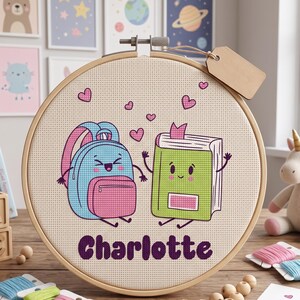May include: A circular wooden embroidery hoop containing a cross-stitch design of a light blue backpack and a green book with faces, surrounded by small pink hearts. The name Charlotte is stitched in purple letters below the characters.