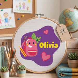 Puede incluir: Un bastidor de bordado de madera circular que contiene tela Aida blanca con un diseño de punto de cruz. El diseño presenta una mochila de dibujos animados rosa con cara, un lápiz amarillo, dos corazones rosas y el nombre Olivia en amarillo sobre un fondo morado.