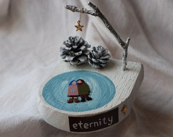 May include: A wooden sculpture with a blue painted circle, two figures in winter clothes, and pine cones. A small branch with a gold star hangs above the circle. The sculpture is painted white and has the word "eternity" written on the bottom.