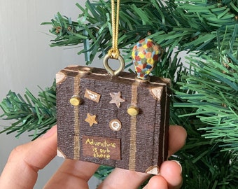 May include: A brown suitcase-shaped Christmas ornament with gold accents and a hot air balloon on top. The ornament has the text "Adventure is out there" on the front.