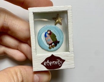 May include: A small white box with a glass front containing a painted scene of two figures holding each other. The figures are painted in blue, brown, and green. A gold star is above the figures. The box has a brown lip with the word "eternity" written in white.