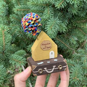 May include: A small wooden house with a painted pine cone tree on top, sitting on a brown suitcase with the words "Adventure is out there!" on the house.