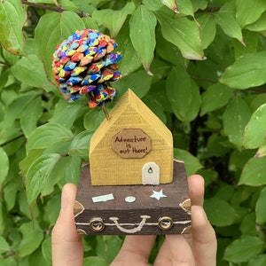 May include: A small wooden house with a yellow exterior and a brown roof sits on top of a brown suitcase. The house has a sign that reads "Adventure is out there!" and a pine cone painted in various colors sits on top of the roof.