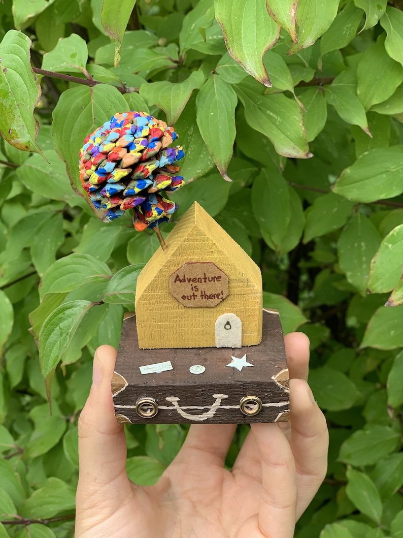 May include: A small wooden house with a yellow exterior and a brown roof sits on top of a brown suitcase. The house has a sign that reads "Adventure is out there!" and a pine cone painted in various colors sits on top of the roof.
