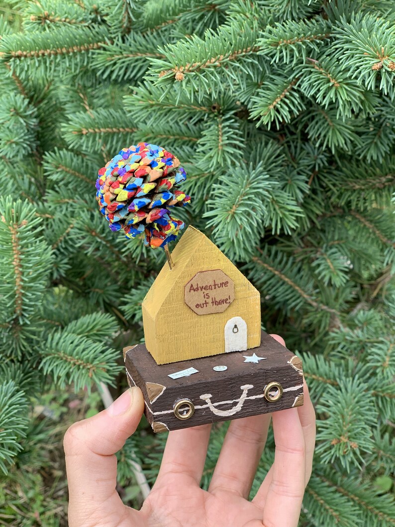May include: A small wooden house with a painted pine cone tree on top, sitting on a brown suitcase with the words "Adventure is out there!" on the house.