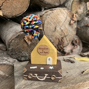 May include: A wooden house with a yellow exterior and a brown roof sits on top of a brown suitcase with a white star and a smiley face. The house has a sign that reads "Adventure is out there!" and a colorful pine cone sits on top of the roof.