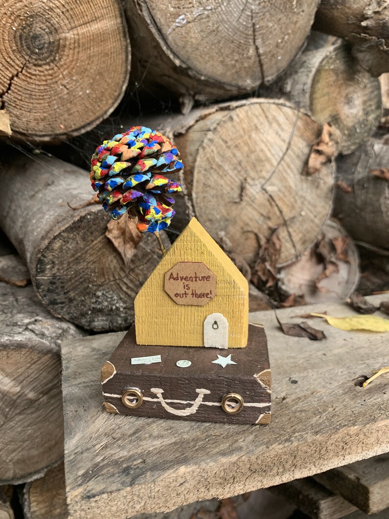 May include: A wooden house with a yellow exterior and a brown roof sits on top of a brown suitcase with a white star and a smiley face. The house has a sign that reads "Adventure is out there!" and a colorful pine cone sits on top of the roof.