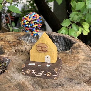 May include: A wooden house with a yellow exterior and a brown suitcase base. The house has a sign that reads "Adventure is out there!" and a pine cone painted in rainbow colors is on top of the house.