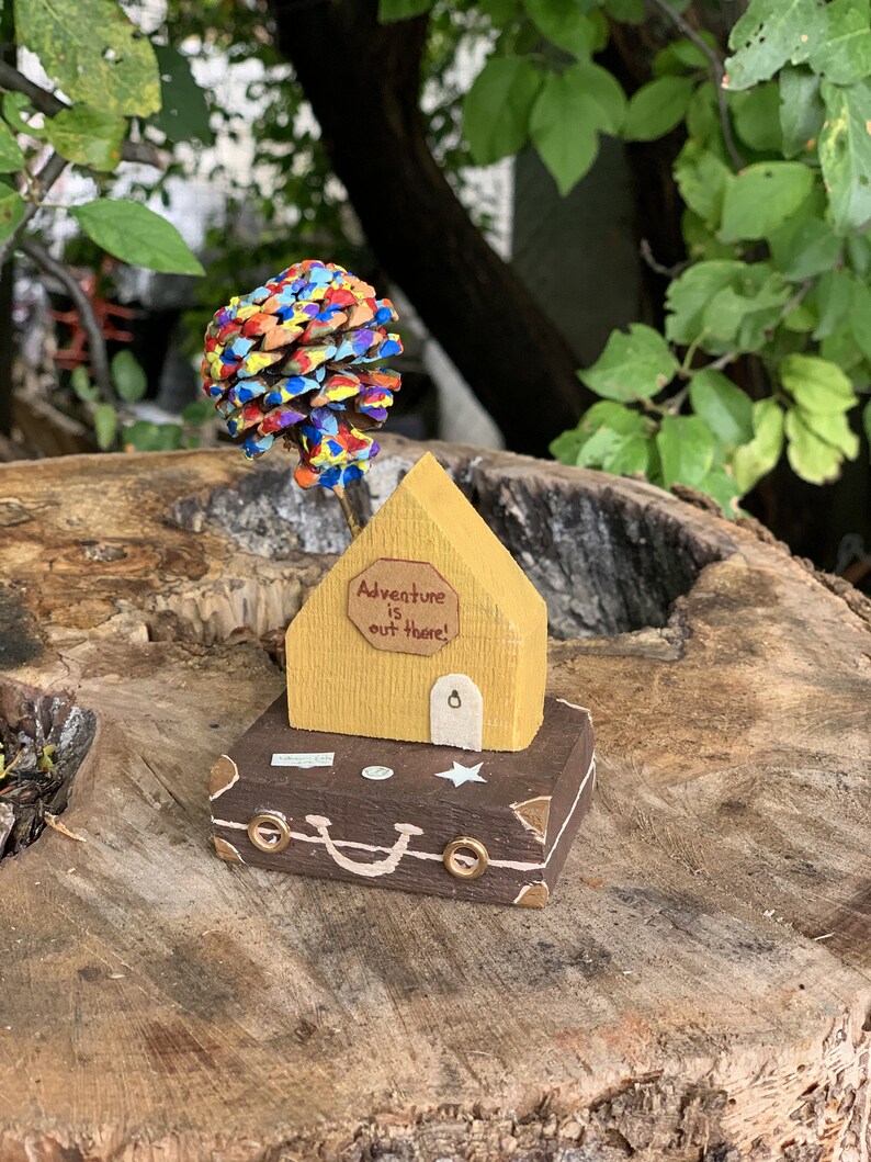 May include: A wooden house with a yellow exterior and a brown suitcase base. The house has a sign that reads "Adventure is out there!" and a pine cone painted in rainbow colors is on top of the house.
