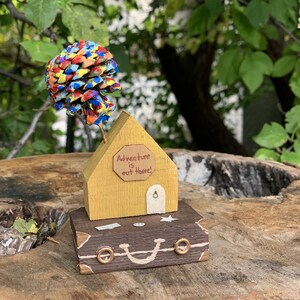 May include: A yellow wooden house with a sign that reads "Adventure is out there!" sits on top of a brown suitcase with gold accents. A colorful pine cone with painted dots sits on top of the house.