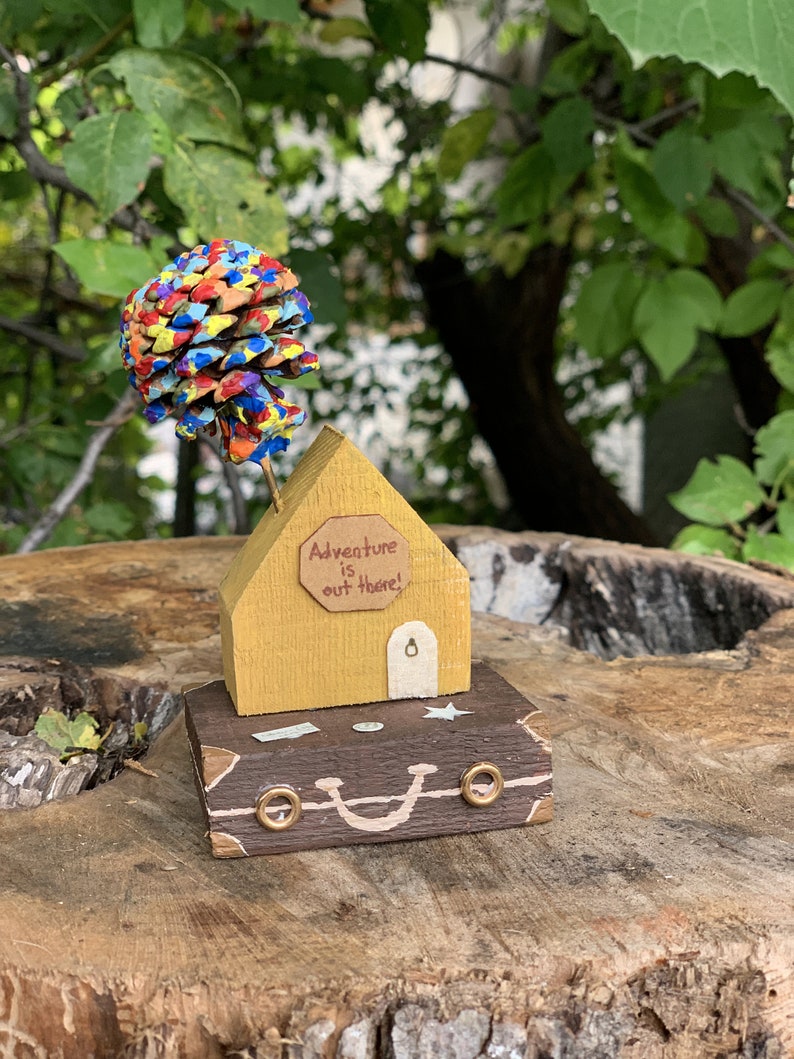 May include: A yellow wooden house with a sign that reads "Adventure is out there!" sits on top of a brown suitcase with gold accents. A colorful pine cone with painted dots sits on top of the house.