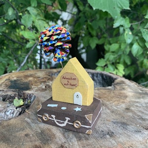 May include: A miniature wooden house with a yellow exterior and a brown suitcase base. The house has a sign that reads "Adventure is out there!" and a pine cone painted in rainbow colors is attached to the roof.