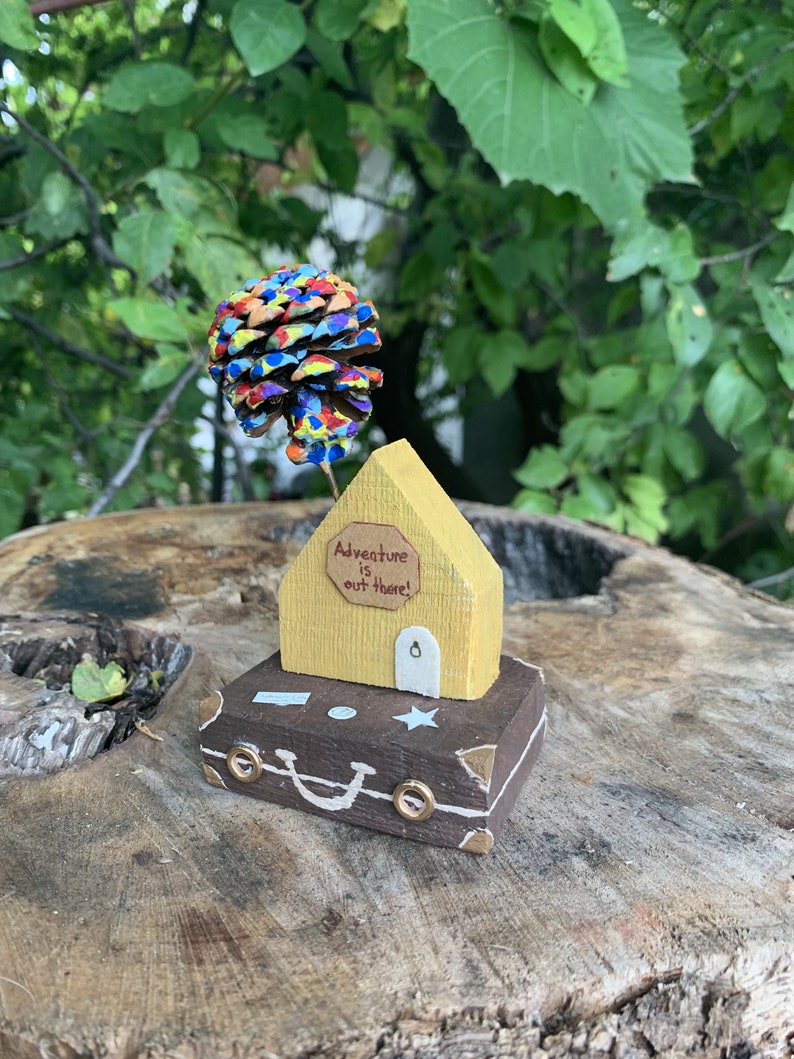 May include: A miniature wooden house with a yellow exterior and a brown suitcase base. The house has a sign that reads "Adventure is out there!" and a pine cone painted in rainbow colors is attached to the roof.