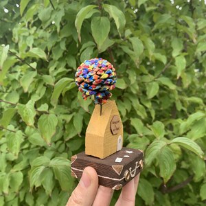 May include: A small wooden house with a colorful pine cone on top, sitting on a wooden suitcase. The house has a door and a sign that says "Adventure is out there".