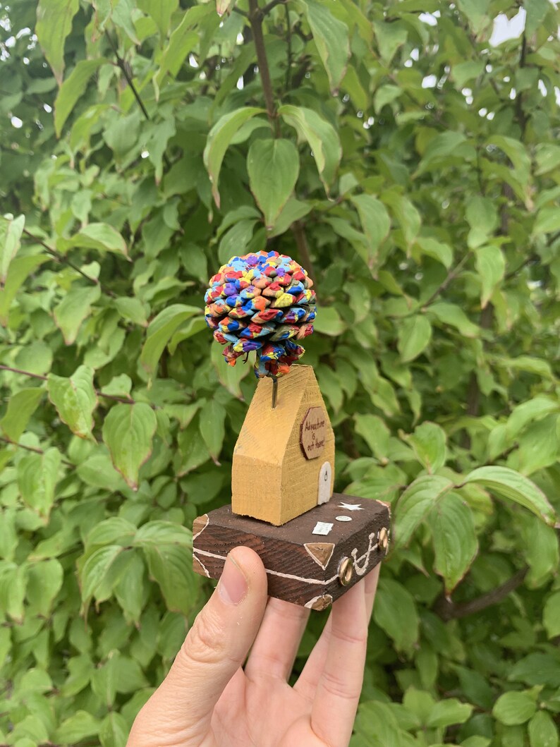 May include: A small wooden house with a colorful pine cone on top, sitting on a wooden suitcase. The house has a door and a sign that says "Adventure is out there".