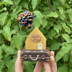 May include: A small wooden house with a yellow exterior and a brown door. The house has a sign that reads "Adventure is out there!" and is sitting on top of a brown suitcase with a painted smile. A colorful pine cone sits on top of the house.