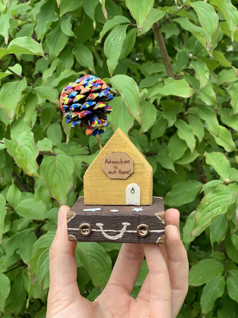 May include: A small wooden house with a yellow exterior and a brown door. The house has a sign that reads "Adventure is out there!" and is sitting on top of a brown suitcase with a painted smile. A colorful pine cone sits on top of the house.