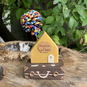 May include: A miniature wooden house painted yellow with a sign that reads "Adventure is out there!"  The house is sitting on top of a brown wooden suitcase with a smiling face painted on it. A colorful pine cone is attached to the top of the house.
