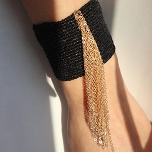 May include: A black cuff bracelet with gold chain fringe. The cuff is made of a textured, woven material. Gold chains cascade from the cuff, creating a stylish accessory.
