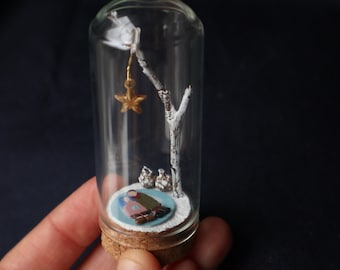 May include: A miniature winter scene inside a glass bottle with a cork stopper. A small, painted figure of a couple sits under a white twig tree with a gold star hanging from a branch. The base of the bottle is covered in white paint to resemble snow.