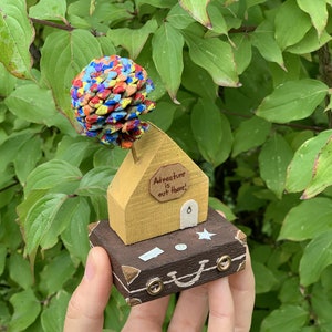 May include: A small wooden house with a yellow exterior and a brown roof sits on top of a brown suitcase. The house has a sign that reads "Adventure is out there!" and a small door. The house is topped with a colorful pine cone.