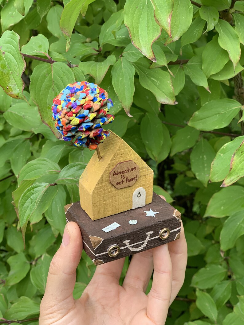 May include: A small wooden house with a yellow exterior and a brown roof sits on top of a brown suitcase. The house has a sign that reads "Adventure is out there!" and a small door. The house is topped with a colorful pine cone.