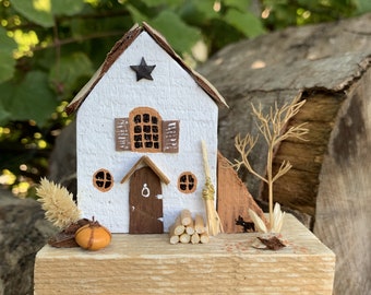 May include: A miniature white house with a brown roof, shutters, and door. A black star decorates the roof. The house sits on a wooden base with a small pumpkin, broom, and firewood. A small tree and a black dog are also present.