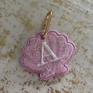 May include: A pink seashell-shaped keychain with a white embroidered letter "A". The keychain has a gold-colored clasp. The seashell design features a floral pattern and a white border.