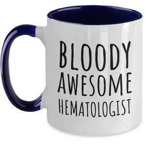 May include: White ceramic coffee mug with a blue rim and handle. The mug has black text that reads "BLOODY AWESOME HEMATOLOGIST".