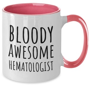 May include: White coffee mug with a pink rim and handle. The mug has black text that reads "BLOODY AWESOME HEMATOLOGIST".