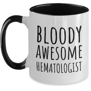 May include: White coffee mug with a black handle and black rim. The mug has black text that reads "BLOODY AWESOME HEMATOLOGIST".